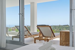 A balcony with two wooden lounge chairs, a bed with white linens, and a view of the coastline. - Marina Retreat by IIM (Estepona)