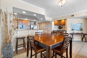 A spacious dining area with a large wooden table and chairs, a kitchen with wooden cabinets, and a decorative fish wall art piece. - Colonnades 302 (Gulf Shores)