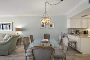 A dining area with a glass table, chairs, and a chandelier. There is a kitchen with white cabinets and a countertop. - Shipwatch D302 (Pensacola)