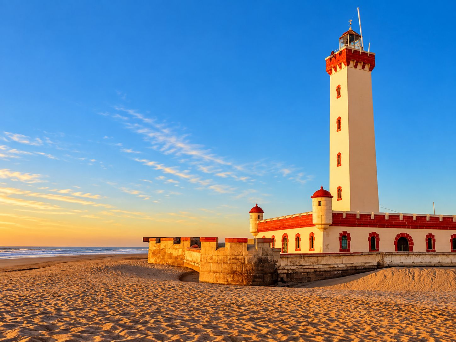 Un faro con techo rojo y paredes blancas, ubicado en una playa arenosa bajo un cielo despejado.