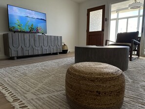 A living room with a television on a wooden cabinet, a round woven ottoman, a large rug, and a view of the outdoors through a door. - 1BR condo on Sapphire Beach - sleeps 4! (St. Thomas)
