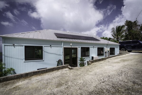 A single-story building with a light blue exterior, a metal roof, and a small front porch. - Magen`s Bay Villa | Tesla Solar Power (St. Thomas)
