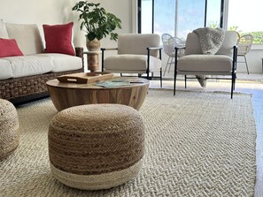 Ein Wohnzimmer mit einem runden Holztisch, einem gewebten Hocker und einem großen Teppich. Zwei Sessel und eine Couch mit Kissen sind ebenfalls vorhanden.