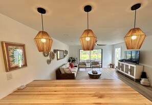 Ein geräumiges Wohnzimmer mit Holzboden, einer Couch, einem Couchtisch und einem Fernseher auf einem Schrank. Drei Pendelleuchten sind an der Decke befestigt.