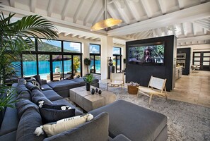 A spacious living area with a large sofa, a coffee table, and a TV. There are plants, a chair, and a view of the ocean. - The BEST Wedding Spot! 2 pools, gym, beachfront & MUCH MORE! (St. Thomas)