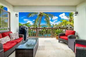 A balcony with wicker furniture, a glass table, and a view of a pool and palm trees. - Amazing Location & Value! A few steps to the beach, walk to the restaurant/bar! (St. Thomas)
