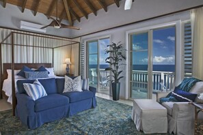 A room with a bed, a sofa, a chair, a large window with a view of the sea, and a ceiling fan. - Luxury Villa with Infinity Pool Near Magens Bay (St. Thomas)