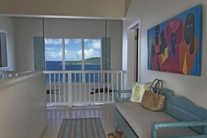 A balcony with a view of the sea, a blue sofa with a woven bag, and a colorful painting on the wall. - Cliff-side 5BR villa - stunning down-island views & 5 mins from Magen`s Bay! (St. Thomas)