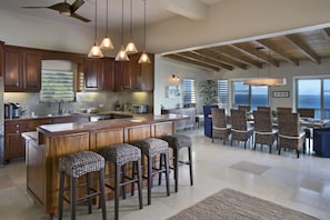 A spacious kitchen with wooden cabinets, a large island with bar stools, pendant lighting, and a view of the ocean through large windows. - Luxury Villa with Infinity Pool Near Magens Bay (St. Thomas)