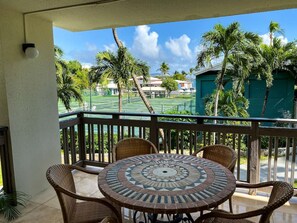 A balcony with wicker chairs and a mosaic table, overlooking a tennis court and palm trees. - 2BR just steps away from the beach and pool (St. Thomas)