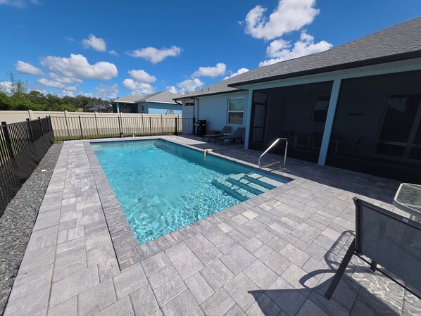 A rectangular swimming pool with a tiled deck, a metal ladder, and a covered patio area with chairs and a table.