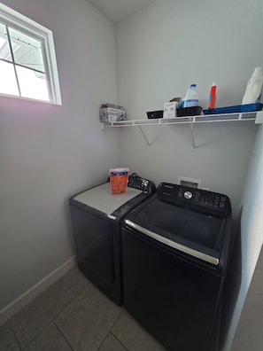 A laundry area with a front-loading washer and dryer, a shelf with cleaning supplies, and a window providing natural light.