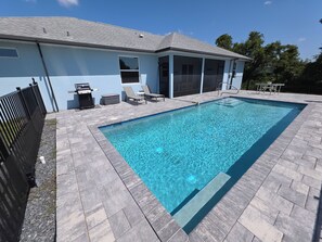 A rectangular swimming pool with a tiled deck, a covered patio area with chairs, and a grill.