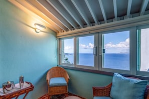Ein Zimmer mit Meerblick, ein Korbstuhl, ein kleiner Tisch mit Tablett und eine Wandleuchte.