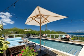 A poolside area with a large umbrella, lounge chairs, and a view of the sea. - 4BR Villa, Private Pool, 360° Ocean Views (St. Thomas)