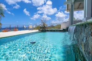 A clear blue pool with a waterfall feature, surrounded by a white deck and chairs, overlooking a scenic view. - Vessup View · 5BR -Walk to the beach! Close to the Ritz Carlton! (St. Thomas)