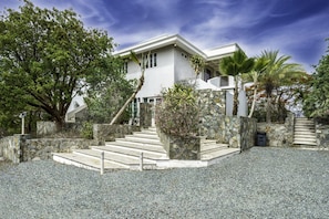 Une maison moderne avec un escalier en pierre menant à l’entrée, entourée d’une végétation luxuriante et sous un ciel dégagé.
