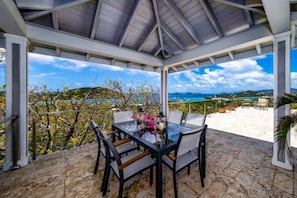 Un espace extérieur abrité, avec une table et des chaises, offrant une vue panoramique sur un paysage verdoyant, avec des arbres et la mer à l’horizon.