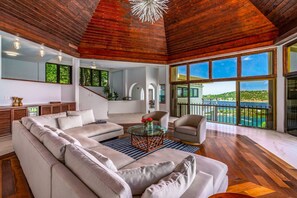 A spacious living room with a large sectional sofa, a glass coffee table, and a view of the ocean through large windows. - 5BR Private Villa, Huge Pool, Ocean Views (St. Thomas)