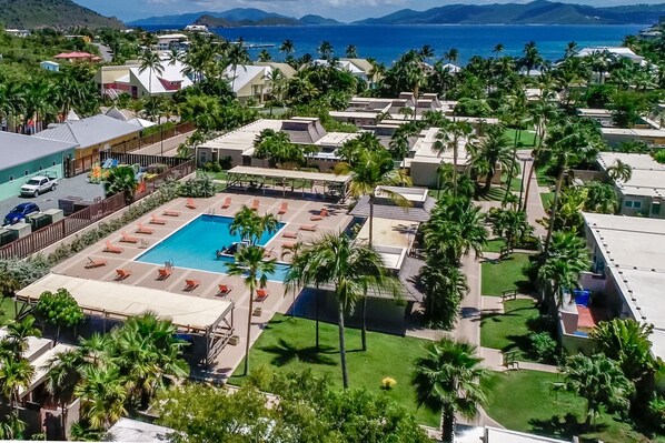 Eine Vogelperspektive zeigt einen Resortkomplex mit einem zentralen Schwimmbecken, umgeben von Liegestühlen, Palmen und Blick auf den Ozean.