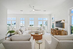 A spacious living room with a large sofa, a coffee table, and a television. - CSB~Ovation~5 BR Luxury Home~Beach, Bay, Pools Access~Panoramic Gulf & Bay Views (Port St. Joe)