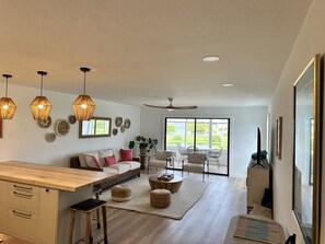 A modern living room with a sofa, coffee table, and chairs. There is a dining area with a wooden table and chairs. The room has large windows and a ceiling fan. - Dual Sapphire Beach Condos w/ Pool (St. Thomas)