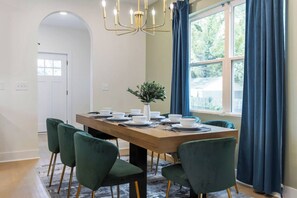 A dining area with a wooden table set for four, green upholstered chairs, a chandelier, and a view through a window with blue curtains. - Emerald Oasis | Hot Tub | Game Room | Sleeps 12 (Columbus)