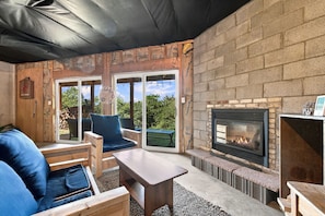 An outdoor patio with a stone fireplace, wooden furniture, and a view through large glass doors.