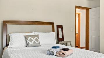 A neatly made bed with pillows and rolled towels, a bedside table with a mirror, and a door leading to another room.