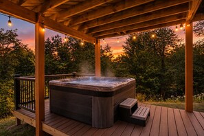 A hot tub under a wooden pergola with string lights, surrounded by trees and a sunset view.
