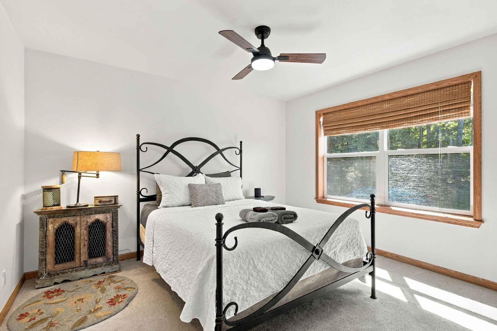 A bedroom with a bed, a nightstand, a lamp, a ceiling fan, and a window with a view of a body of water.