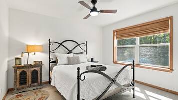 A bedroom with a bed, a nightstand, a lamp, a ceiling fan, and a window with a view of a body of water.