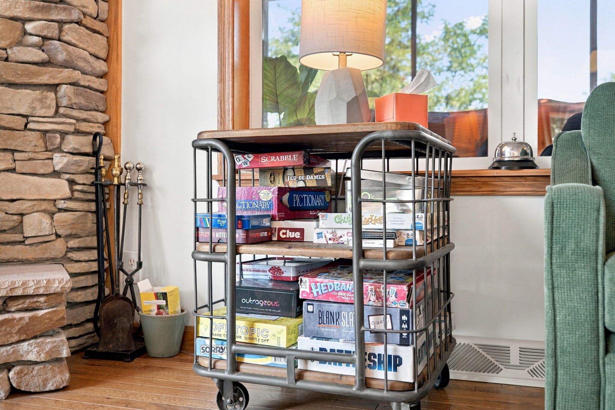 A metal game cart filled with board games in a room with a stone wall and a window.