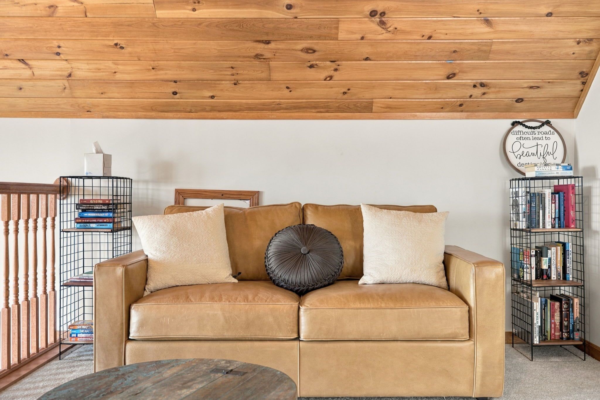 A tan leather sofa with two pillows, a round accent pillow, a metal bookshelf, and a wooden ceiling.
