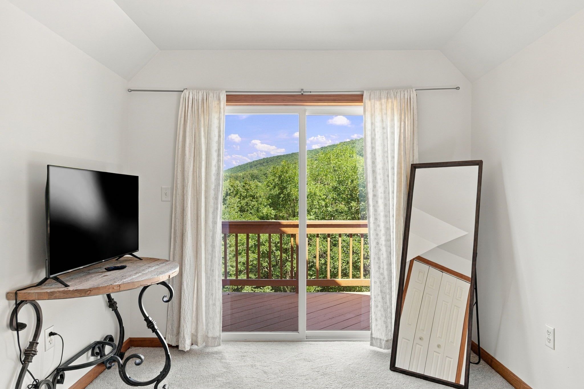 A room with a TV on a wooden stand, a large sliding glass door leading to a balcony, and a white wardrobe.