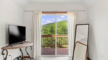 A room with a TV on a wooden stand, a large sliding glass door leading to a balcony, and a white wardrobe.