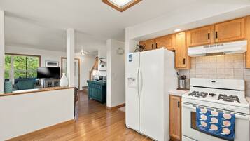 A modern kitchen with wooden cabinets, a white refrigerator, a stove, and a dining area with a television and a teal chair.