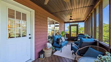 Een overdekt terras met een houten vloer, een witte deur en een zithoek met blauwe kussens en rieten meubels.