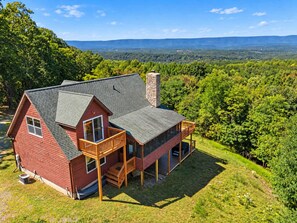 A two-story house with a balcony, surrounded by lush greenery and a view of distant hills.