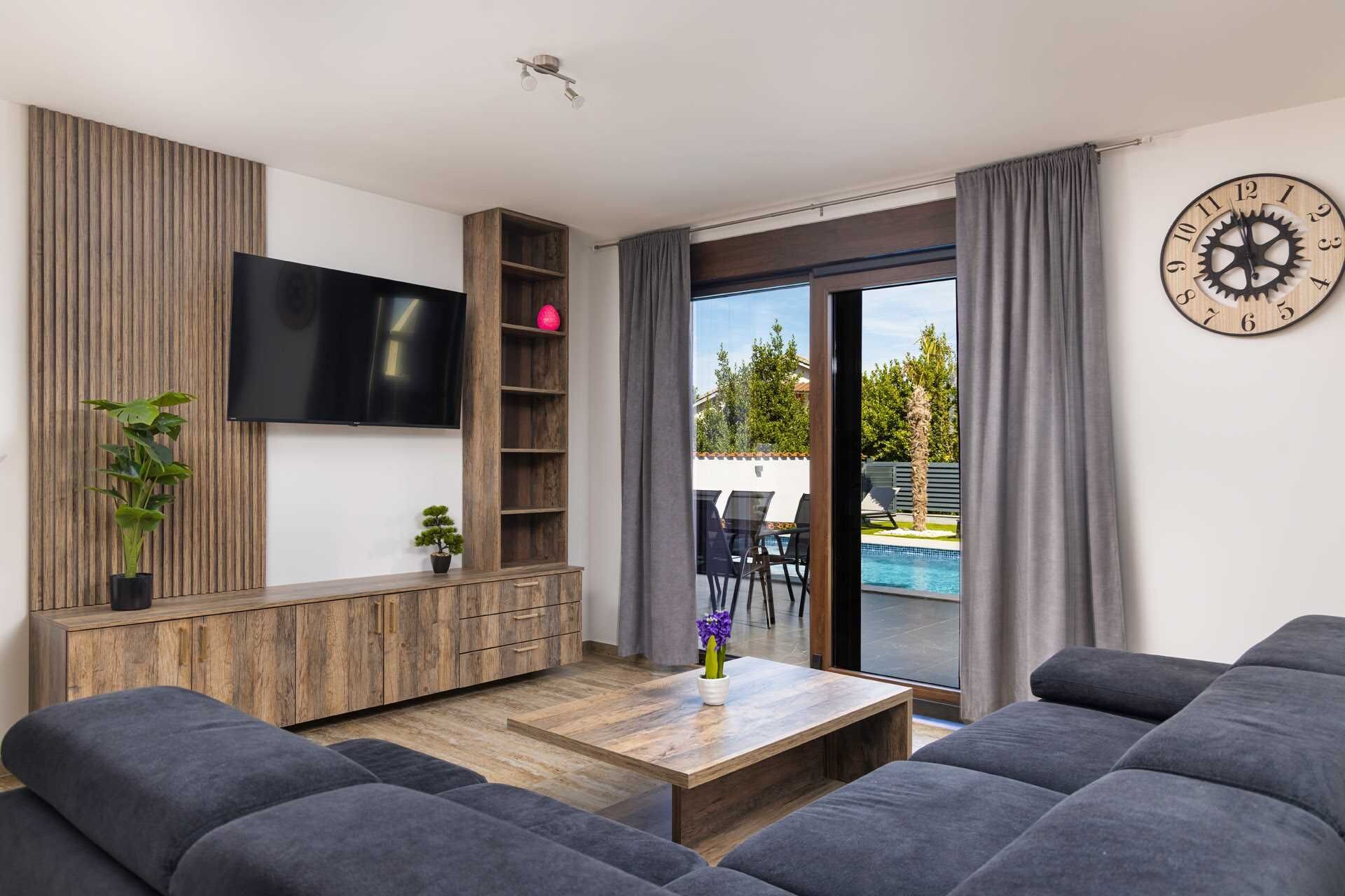 Een moderne woonkamer met een flatscreen televisie, houten schapenkasten, een salontafel en een groot raam met uitzicht op een zwembad en tuinmeubilair.
