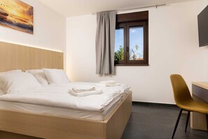 Quarto de hotel com cama, escrivaninha, cadeira, janela com vista e um quadro na parede.