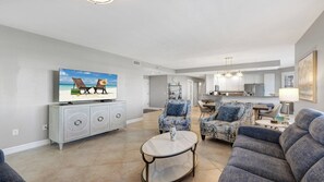 Amplia sala de estar con televisor, dos sillones, un sofá y un comedor con una cocina al fondo.