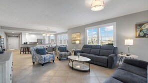 Amplia sala de estar con un sofá gris, sillones estampados y una mesa de centro. Se ve una cocina con armarios blancos y un comedor con mesa y sillas al fondo.