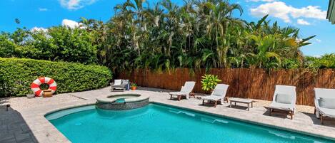 A swimming pool with a wooden deck, surrounded by palm trees and a bamboo fence.