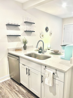 A kitchen with a double sink, white cabinets, and a dishwasher. There are shelves above the sink with glassware and a potted plant on the counter.
