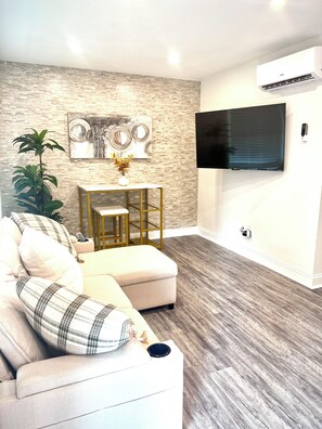 A living room with a sectional sofa, a coffee table, a wall-mounted TV, and an air conditioning unit. - NoDa Cottage Near Uptown (Charlotte)