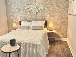 A modern bedroom with a patterned wall, a bed with white bedding, two bedside lamps, and a small round table with a speaker and a smartphone. - NoDa Cottage Near Uptown (Charlotte)
