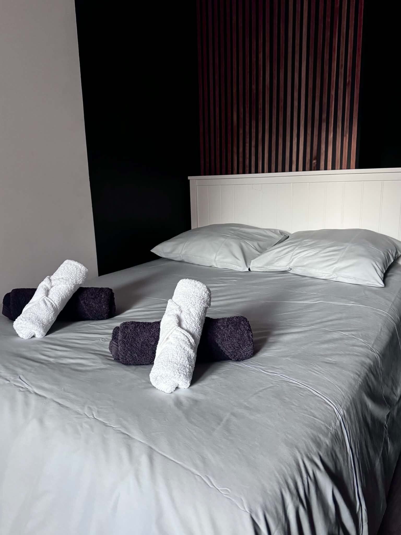 Ein ordentlich bezogenes Bett mit grauen Bettwäsche und zwei zusammengerollten Handtüchern darauf.