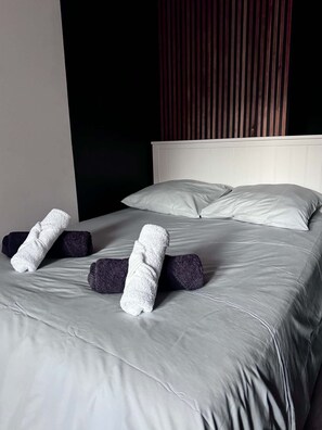 A neatly made bed with grey linens and two rolled towels placed on the bed.