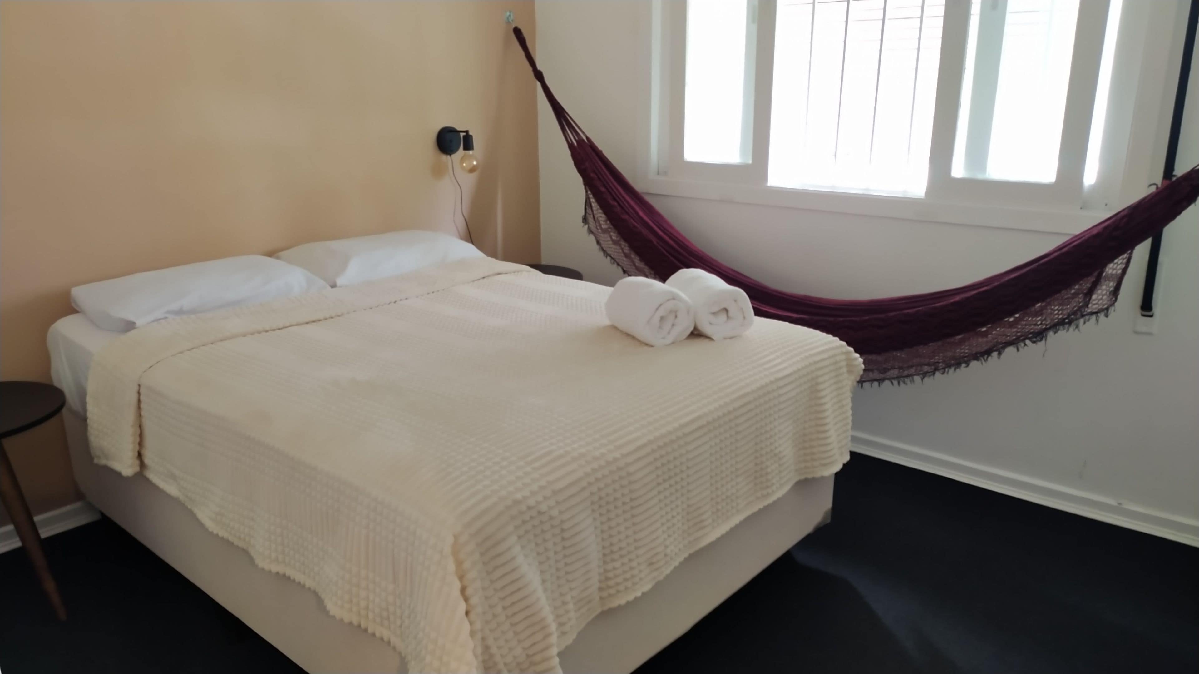 Ein Bett mit weißer Decke und Kissen, ein kleiner Nachttisch und eine Hängematte an der Wand.